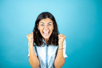 Fototapeta premium Young beautiful woman wearing a denim jumpsuit over isolated blue background very happy and excited making winner gesture with raised arms, smiling and screaming for success.