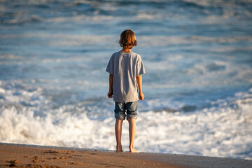 Boy standing on sand looking out at rough water. Late afternoon sunlight