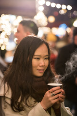 Asian girl holding her cup of coffee with both hands before drinking it in Princes Street Gardens, Edinburgh, UK.