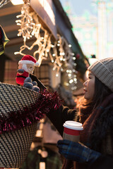 An Asian girl wearing a woolly hat holds a small Santa toy on her hand and smiles in front of a market stall in Edinburgh City center, Scotland, with Christmas decorations on the background.