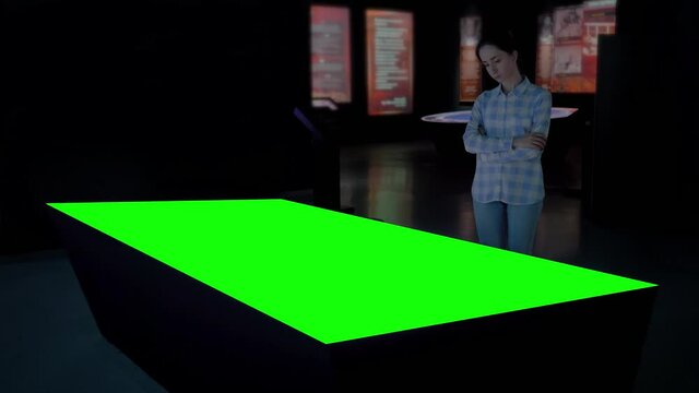 Green screen, mockup, science, copyspace, template, chroma key, technology concept. Woman looking at blank green display of interactive table kiosk in dark room of modern exhibition or museum