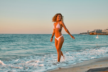 Attractive Caucasian woman in bikini swimsuit strolls on the beach at sunrise enjoying the sound of the waves.