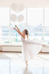 Cute little ballerina in white dress and pointe shoes with white balloons is dancing in the room.