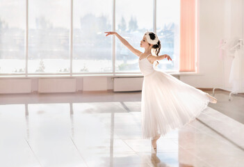 Cute little ballerina in white ballet costume and pointe shoes is dancing in the room.