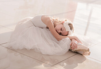 Cute little ballerina in white ballet costume and pointe shoes is posing on floor.