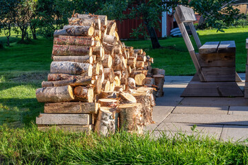 Close up photo of fresh stacked fire wood logs on barbeque spot, garden. A lot of birch logs harvested to be used for grilling. Summer evening, green grass. Blurry apple tree and small wooden cabin - Powered by Adobe