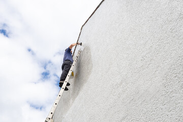 Hardworking man high up a ladder paints side of house a fresh white colour.