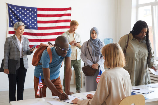 Large Multi-ethnic Group Of People Registering At Polling Station Decorated With American Flags On Election Day, Copy Space