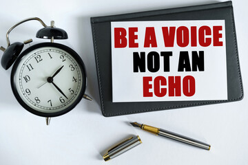 On the table there is a clock, a pen, a notebook and a card on which the text is written - BE A VOICE NOT AN ECHO