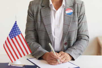 Cropped portrait of mature woman sitting at desk with American flag at polling station on election...