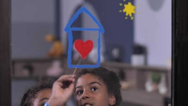 Cheerful dark-skinned curly sisters painting red heart inside house on window glass during lockdown, close-up of paint drawing, symbolizing safety, need to stay at home for coronavirus prevention