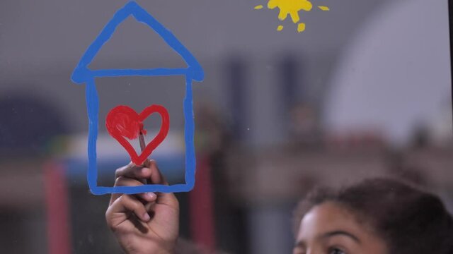Close-up of cute smiling mixed race schoolgirls enjoying painting on window glass. Little artists creating soulful picture with heart inside house, symbolizing safety at home during 2019-ncov pandemic