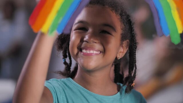 Close-up of cute dark-skinned girl painting hope symbol - multicolored rainbow on window glass. Happy mixed race preteen schoolgirl enjoying symbolic art while staying at home during covid lockdown