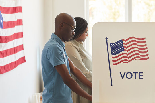 Waist up side view portrait of young African-American people standing in voting booth decorated with USA flags, copy space