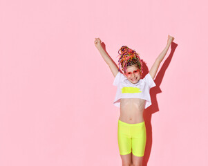 Excited teenage girl with African braids, in sunglasses, yellow shorts and a white top. She raised her hands and, rejoicing or shouting, poses against a pink studio background.