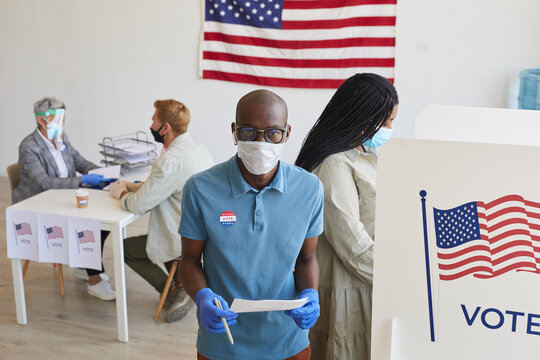 High angle portrait of young African-American voter wearing mask standing by booth and looking at camera on post-pandemic election day, copy space