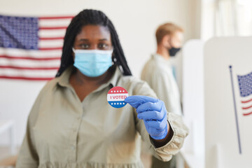Blurred portrait of African-American woman holding I VOTED sticker while standing t polling station...