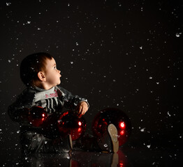 Baby girl in gray boots and sparkling suit. She posing with three red balls, sitting on floor. Twilight black background. Close up