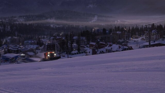 snow truck with glowing headlights cleans mountain slope at ski resort against distant town and forest in cold winter evening