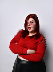 Overweight red-haired woman in a red studded top, black leather skirt and earrings. She stands with her arms folded in a twisted pose