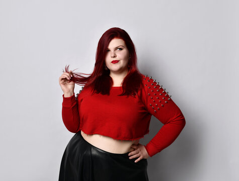 Obese redhead lady in red spiked top, black leather skirt, earrings. Touching face, put hand on waist, posing isolated on white
