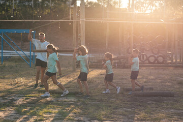Male fitness coach instructing while kids carrying logs at a bootcamp
