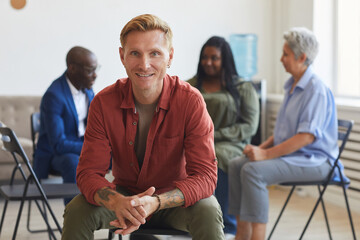 Portrait of smiling tattooed man looking at camera during support group meeting with people in background, copy space