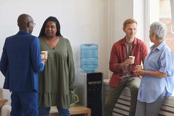 Multi-ethnic group of people drinking coffee and chatting while standing by water cooler in support meeting, copy space - Powered by Adobe