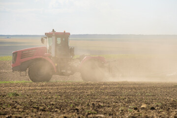 A red farm tractor in a cloud of dust cultivates the soil in the field with a cultivator after...