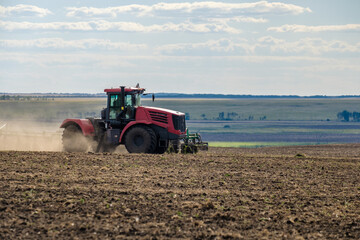 A red farm tractor in a cloud of dust cultivates the soil in the field with a cultivator after harvest. Summer sunny day. Fertile land. Modern agricultural machinery. Copy space. High quality photo