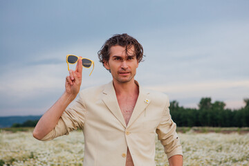 Tall handsome man standing in camomile flowers field and posing with yellow sunglasses