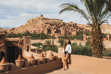 Beautiful young woman happy to travel in Morocco. Ait-Ben-Haddou kasbah on background. Wearing in white shirt and jeans shorts.