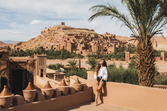 Beautiful Young Woman Happy To Travel In Morocco. Ait-Ben-Haddou Kasbah On Background. Wearing In White Shirt And Jeans Shorts.