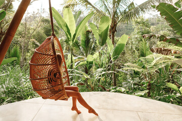 Back view yourist woman swing on wicker rattan hang chair in the jungle, nature view. Enjoy vacation on tropical Bali island