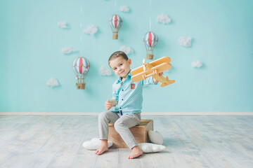 A boy plays with a wooden airplane against the background of a decorative blue wall with clouds and hot air balloons. A pilot's dream.