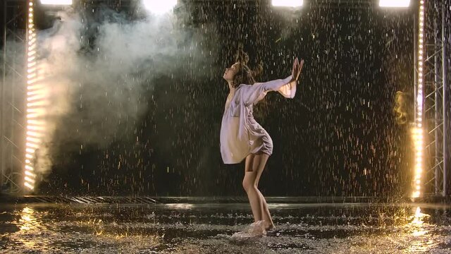 A woman in a white shirt is dancing on the stage the dramatic dances of modern ballet in the rain. A ballet dancer moves plastic performing modern choreography in smoke. Slow motion.