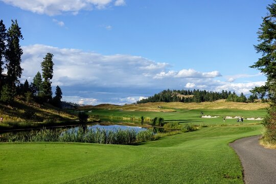 A beautiful view of a par 4 dog leg left from a tee box over a lake.  It is a sunny day with forest in the background, and bunkers lining the fairway.