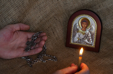 Male Hands hold a pectoral cross and a burning candle in front of a Christian icon.