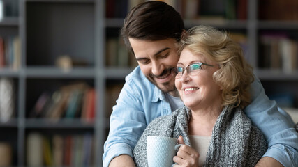 Smiling young man hugging mature mother drinking tea or coffee, smiling middle aged woman holding...
