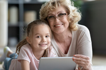 Head shot portrait close up smiling mature grandmother wearing glasses and granddaughter using tablet looking at camera, lying on warm floor, enjoying leisure time with electronic device together