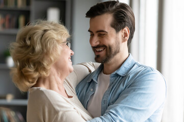 Close up smiling mature mother and adult son hugging, standing in living room at home, family enjoying tender moment, happy overjoyed beautiful middle aged woman and young man cuddling