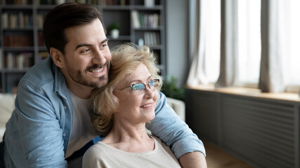 Close up dreamy young man and mature woman hugging, looking to aside, sitting in modern living...