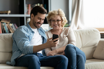 Overjoyed mature woman wearing glasses and adult son using smartphone together, looking at screen,...