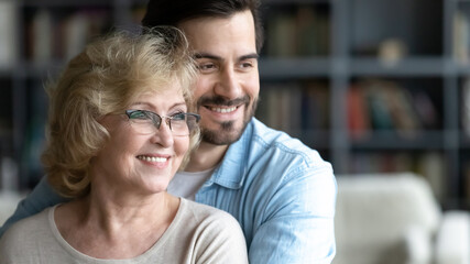 Close up smiling young man and mature mother wearing glasses dreaming together, happy dreamy middle aged woman and adult son hugging, looking to aside, dreaming about good future, standing at home