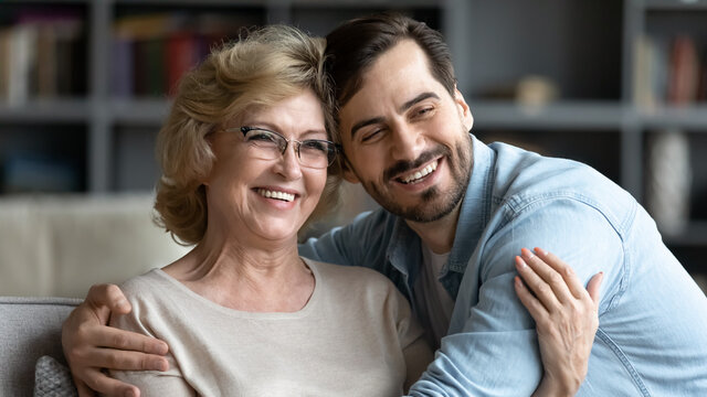 Overjoyed mature woman wearing glasses and adult son hugging, posing for photo, hugging, sitting on couch in living room, excited happy middle aged mother and young man embracing, two generations