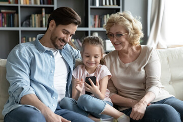 Little smiling girl with father and mature grandmother wearing glasses using smartphone together, looking at screen, watching funny video in social network, shopping online, sitting on couch