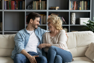 Happy mature woman wearing glasses and young man chatting, talking sitting on couch in living room,...