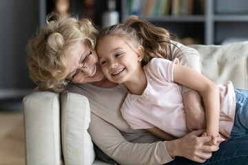 Laughing mature grandmother playing with pretty little granddaughter on couch, lying on cozy sofa, hugging, happy middle aged woman wearing glasses and cute girl enjoying leisure time at home