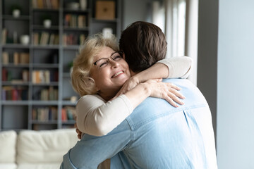 Close up smiling mature woman wearing glasses hugging adult son, standing in living room at home,...