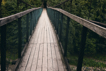 Fototapeta premium Landscape view of Long Steel Suspension bridge. Old small wooden bridge across river only for pedestrian. Mountain river surrounded by mountains and alpine forest